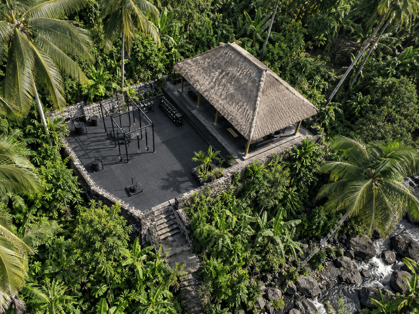 Vue aérienne du plateau d’entraînement noir entouré d’un mur en pierre, avec un rack, des disques, un traîneau et le pavillon au toit de chaume dans la jungle